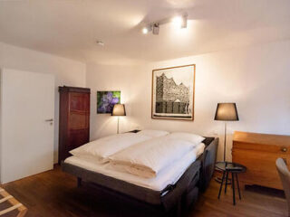 A bedroom features a double bed with white bedding, two black bedside lamps, a wooden wardrobe, a small round table, and wall art. The room has wooden flooring and white walls.