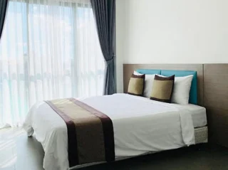 A modern bedroom with a neatly made bed featuring brown accent pillows and a decorative runner, next to large windows with white curtains and gray drapes.