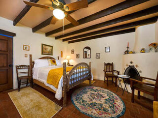 A cozy bedroom with a wooden bed, yellow blanket, ceiling fan, exposed beams, various artworks on the wall, chairs, a small rug, and a fireplace.