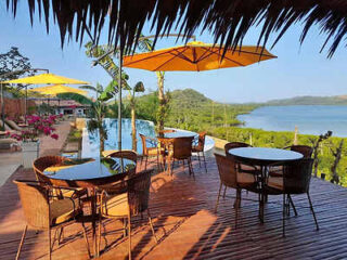 Outdoor seating area with wicker tables and chairs under yellow umbrellas on a wooden deck overlooking an infinity pool and lush green landscape extending to a body of water in the background.