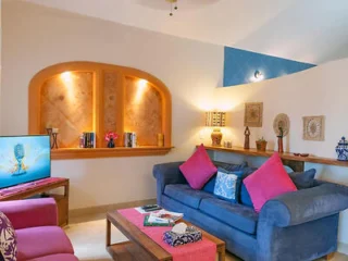 A cozy living room with a blue sofa, pink cushions, a wall-mounted TV, and a wooden coffee table. The wall features two niches with decorative items and a shelf with more decor and plants.