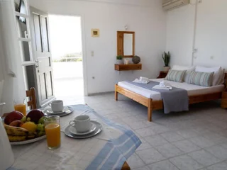 A well-lit room with a made bed, two cups and saucers on a table, a fruit bowl, and a glass of juice. The door is open, revealing an outside view.