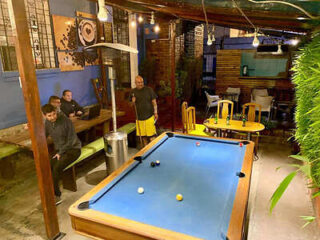 A group of people is gathered in an outdoor area with a pool table. Some are seated at a table while one person stands next to the pool table, holding a cue stick. The area is lit with hanging lights.