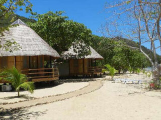 Two thatched-roof huts surrounded by trees on a sandy beach under a clear blue sky. A winding path leads to the huts. White loungers are visible in the distance near the shoreline.