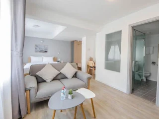 A modern hotel room with a bed, couch, table, desk, and chair. The room has a bathroom with a glass partition and light wood flooring. Bottled water and a small plant are on the table.
