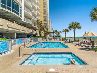 Seaside resort outdoor pool area with palm trees, people swimming, sunbathing on lounge chairs, and tall buildings in the background under a clear blue sky.