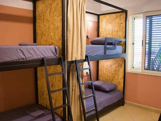 A cozy hostel room with two bunk beds featuring wood-paneled frames and privacy curtains. The room has peach-colored walls and a window with shutters partially open.