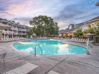 A large round swimming pool surrounded by lounge chairs, with a backdrop of residential buildings and a vibrant sky.