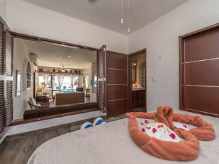 Cozy bedroom with a heart-shaped towel arrangement on the bed, wooden shutters opening to a living area, and a ceiling fan. Living area has large windows, allowing natural light to enter.