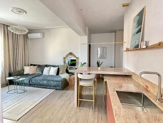 A modern open-plan living area featuring a grey sectional sofa, a marble-topped kitchen island, and minimalist decor including a large mirror and framed art.