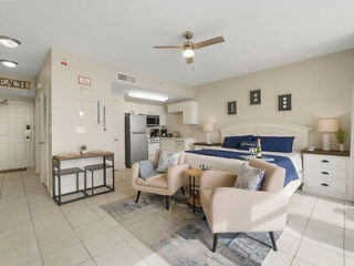 A modern studio apartment with a king-size bed, kitchenette, seating area with two beige chairs, a small table, and a ceiling fan. The room features neutral tones and minimalistic decor.