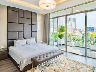A modern bedroom with a large bed, gray padded headboard, a bench at the foot, and a plush rug. The room has floor-to-ceiling windows and a balcony with city views.