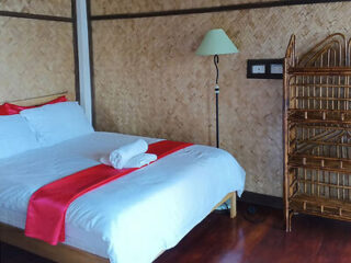 A neatly made bed with a red runner and two folded towels is next to a wicker shelving unit and a floor lamp in a room with woven bamboo walls.