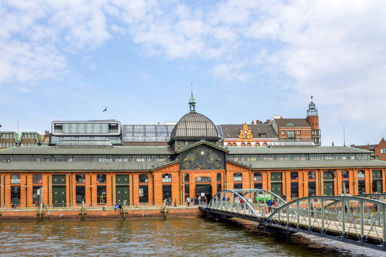 A historic building with a central dome and large windows sits along a waterfront, with a metal pedestrian bridge in the foreground. The sky is partly cloudy.