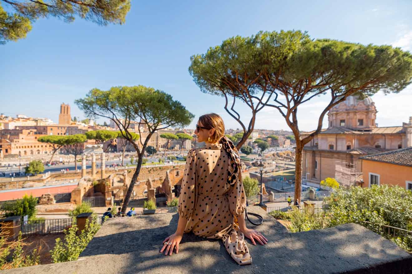 A woman in a polka dot dressW sits on a ledge overlooking the ancient ruins of the Roman Forum, surrounded by lush trees and historic buildings under a clear blue sky