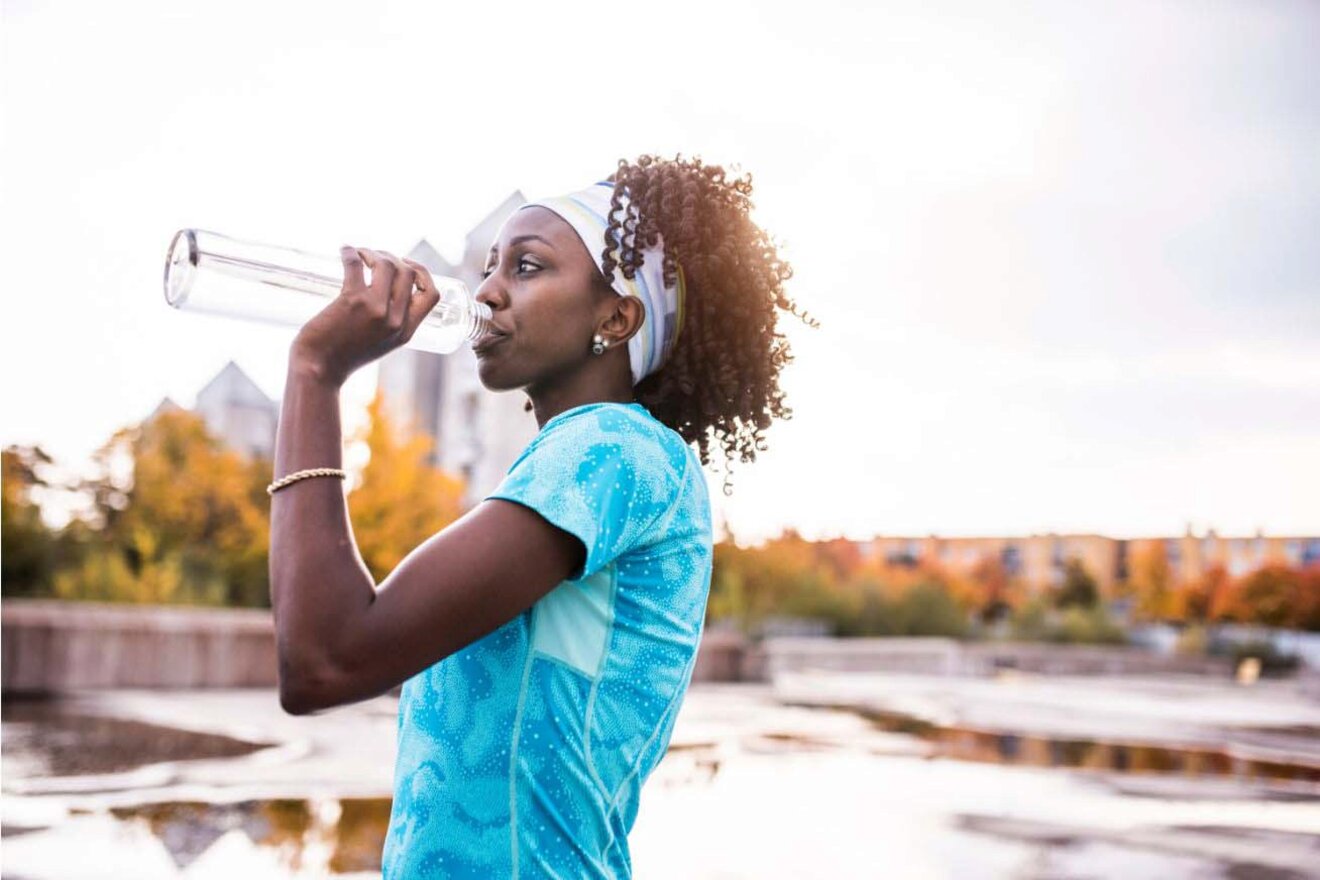 A woman in sportswear drinks water from a bottle outdoors.