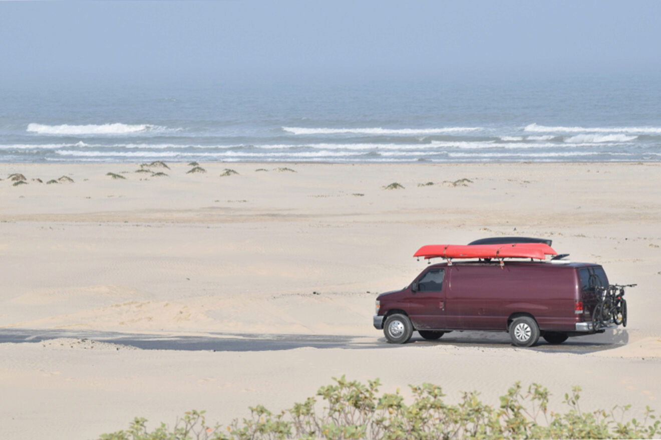 A maroon van parked on a beach with two red kayaks secured on its roof. The ocean and waves are visible in the background, with some shrubs in the foreground.
