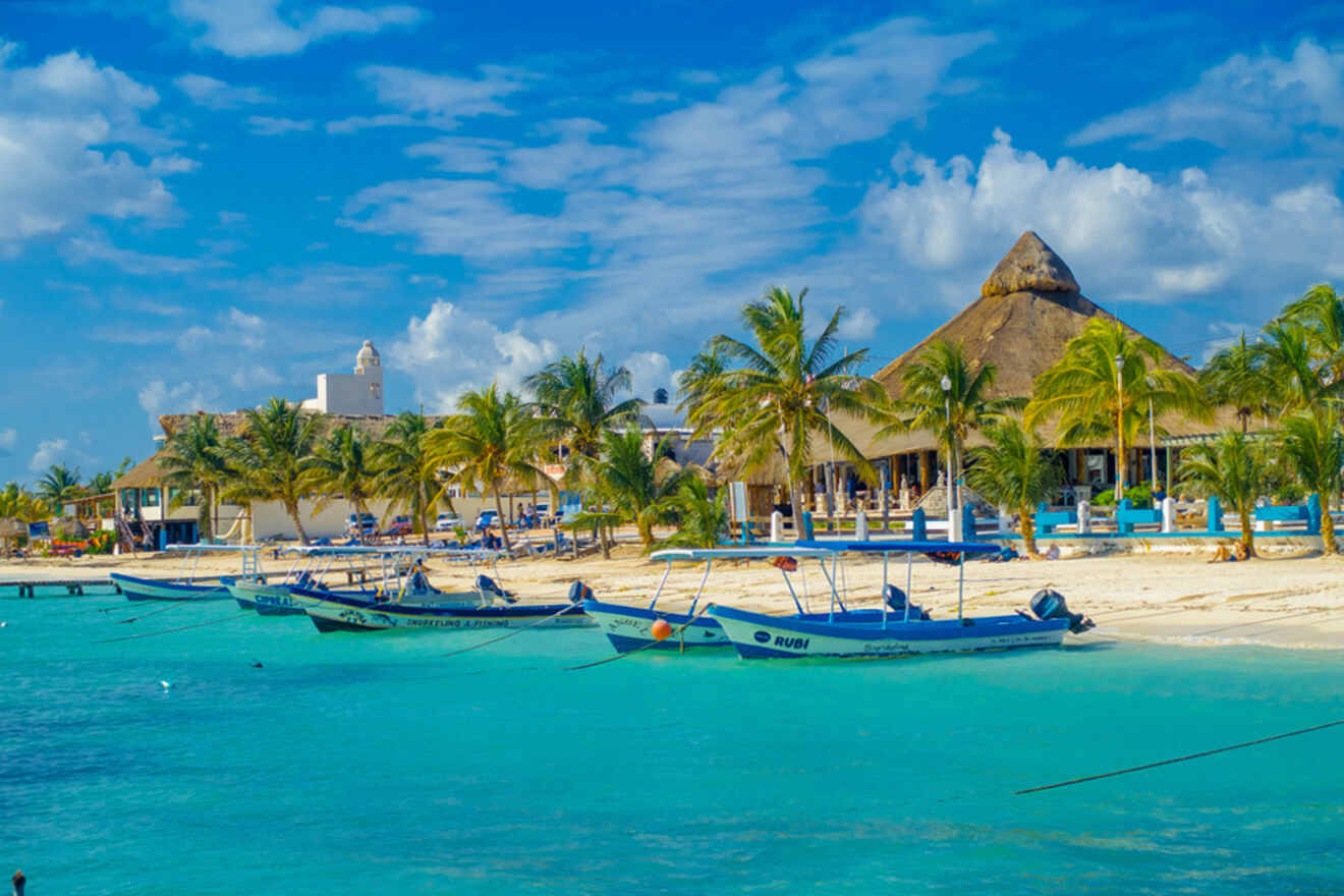 Boats are anchored in turquoise water near a sandy beach with palm trees and buildings under a bright blue sky with some clouds.