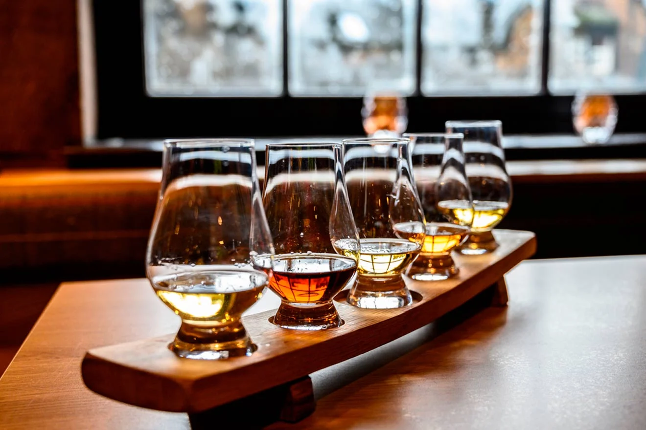 A wooden tray holds five glasses of whiskey, with various hues, placed on a table with a blurred window in the background.