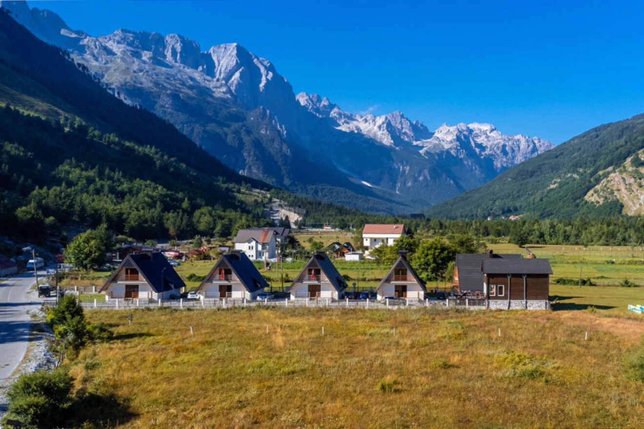 A row of small houses with triangular roofs stands in a grassy field amidst a mountainous landscape, with tall peaks under a clear blue sky in the background.