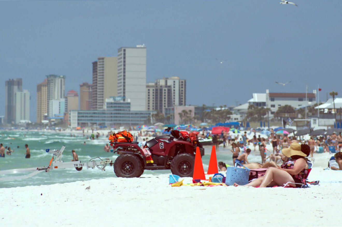 Crowded beach scene with people sunbathing, a lifeguard ATV parked nearby, high-rise buildings in the background, and seagulls flying overhead.