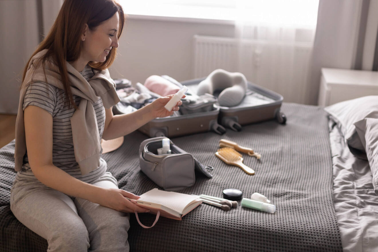 A woman sits on a bed looking at her phone, with an open notebook in her lap. An open suitcase and various personal items are spread out on the bed.