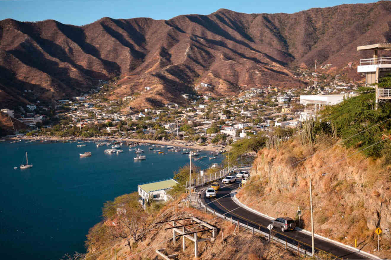 View from a hillside overlooking a coastal town with a bay, boats, and houses nestled in a valley surrounded by arid, brown hills. A winding road with cars is visible in the foreground.