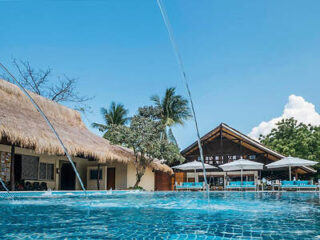 A clear blue pool with water fountains in front of thatched and wooden buildings on a sunny day, surrounded by palm trees.