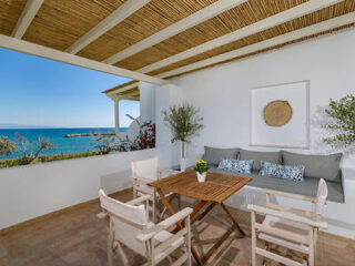 A covered patio featuring a wooden table with chairs, a cushioned bench with pillows, and a decorative wall piece. The patio overlooks the ocean and is roofed with woven material.