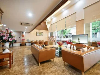 Spacious living area with marble flooring, two brown leather sofas, a large TV, dining table, and large windows letting in natural light. Decor includes flowers and artwork on the walls.