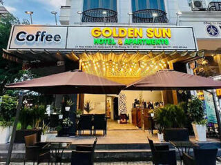 Front view of Golden Sun Hotel & Apartments, featuring outdoor seating under umbrellas with a large signboard for "Coffee" on the left. The entrance is adorned with string lights.