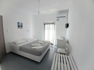 A minimalist white bedroom with a double bed, gray bedspread, and folded towels. There is a window with sheer curtains, an air conditioner, a small wall-mounted TV, and a desk with a chair.