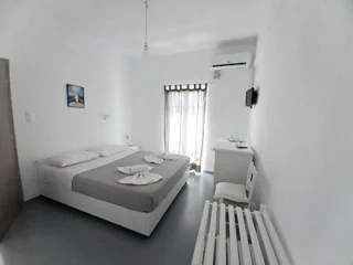 A minimalist white bedroom with a double bed, gray bedspread, and folded towels. There is a window with sheer curtains, an air conditioner, a small wall-mounted TV, and a desk with a chair.