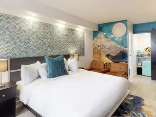 A hotel room features a large bed with white linens and green pillows, a brown sofa, patterned wallpaper, and a mural of a mountain and moon on the back wall near a small kitchenette.