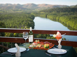 Outdoor dining setup with a table for two featuring a meal, wine bottle, and dessert. The table overlooks a scenic river and hilly landscape in the background.