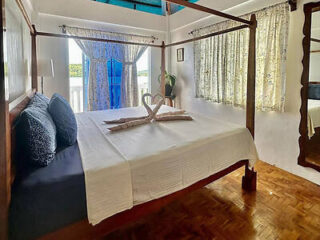 A neatly made four-poster bed with white linens, featuring towel swans on top. The room has a large window with sheer and floral curtains, and a full-length mirror on the right wall.