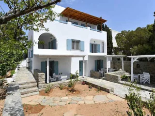 A white two-story house with blue shutters and balconies, surrounded by trees and a stone-paved yard. The house has outdoor seating and a wooden pergola on the upper level.