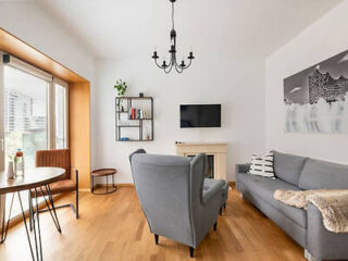 A bright, modern living room with wooden floors, a gray sofa, an armchair, a wall-mounted TV, a round table with two chairs, a small bookshelf, and a black chandelier.