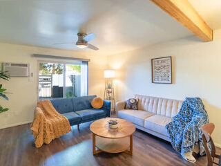 A living room with two couches, one blue and one beige, a ceiling fan, a round wooden coffee table, a floor lamp, an air conditioner, and a wall-mounted framed artwork.