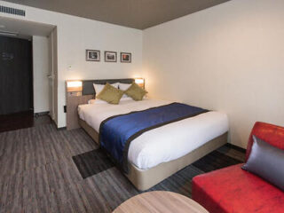 A hotel room with a large bed, blue bed runner, and a red chair, offering a comfortable and modern stay.