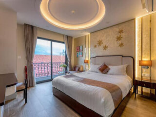 A well-lit hotel room with a large bed, wall-mounted decorations, a desk, and a balcony overlooking red-tiled roofs.