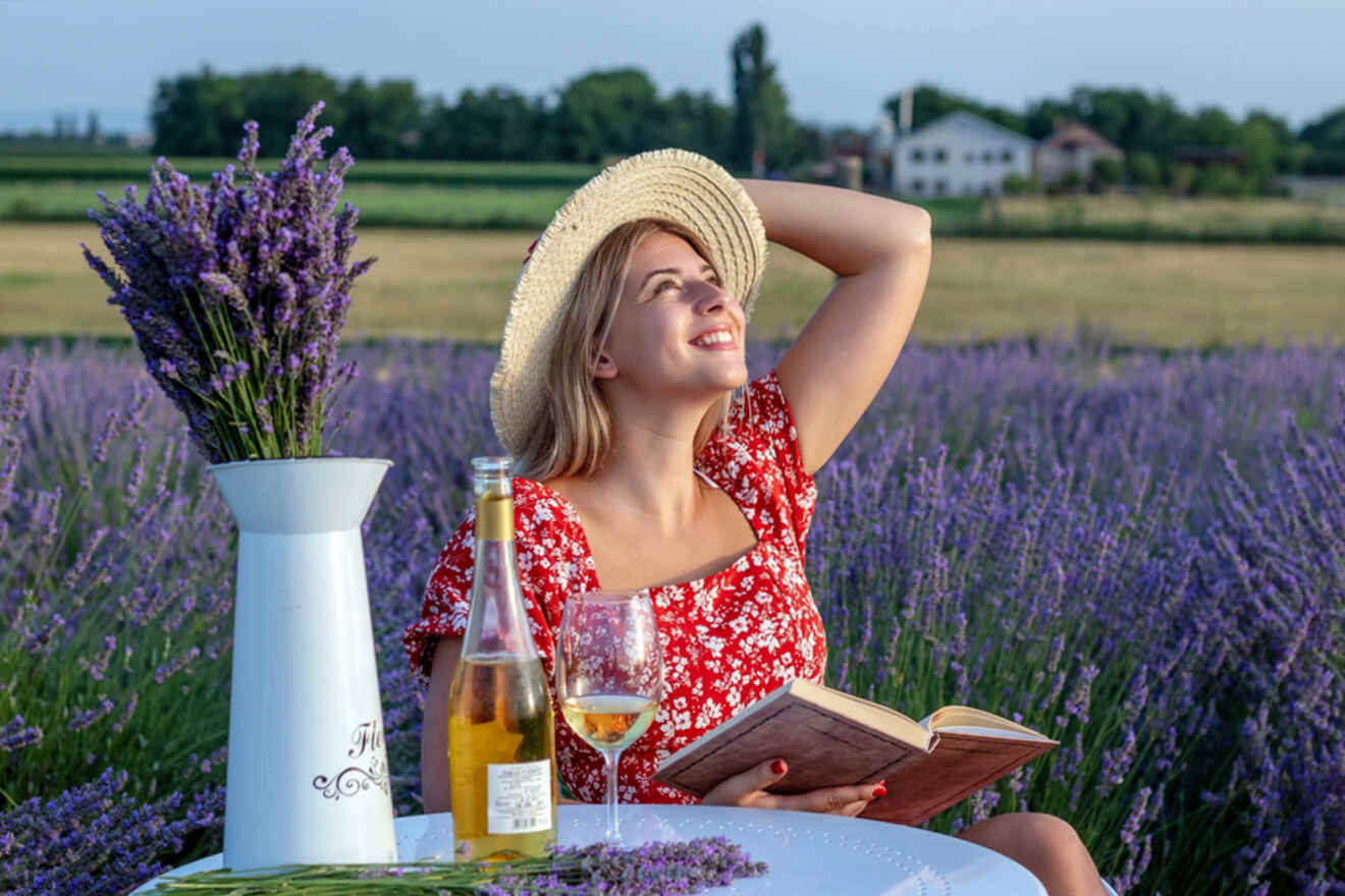 Woman in a red dress and straw hat sits at a table in a lavender field, holding an open book. A wine bottle, glass, and lavender bouquet are on the table. She gazes upward, smiling.