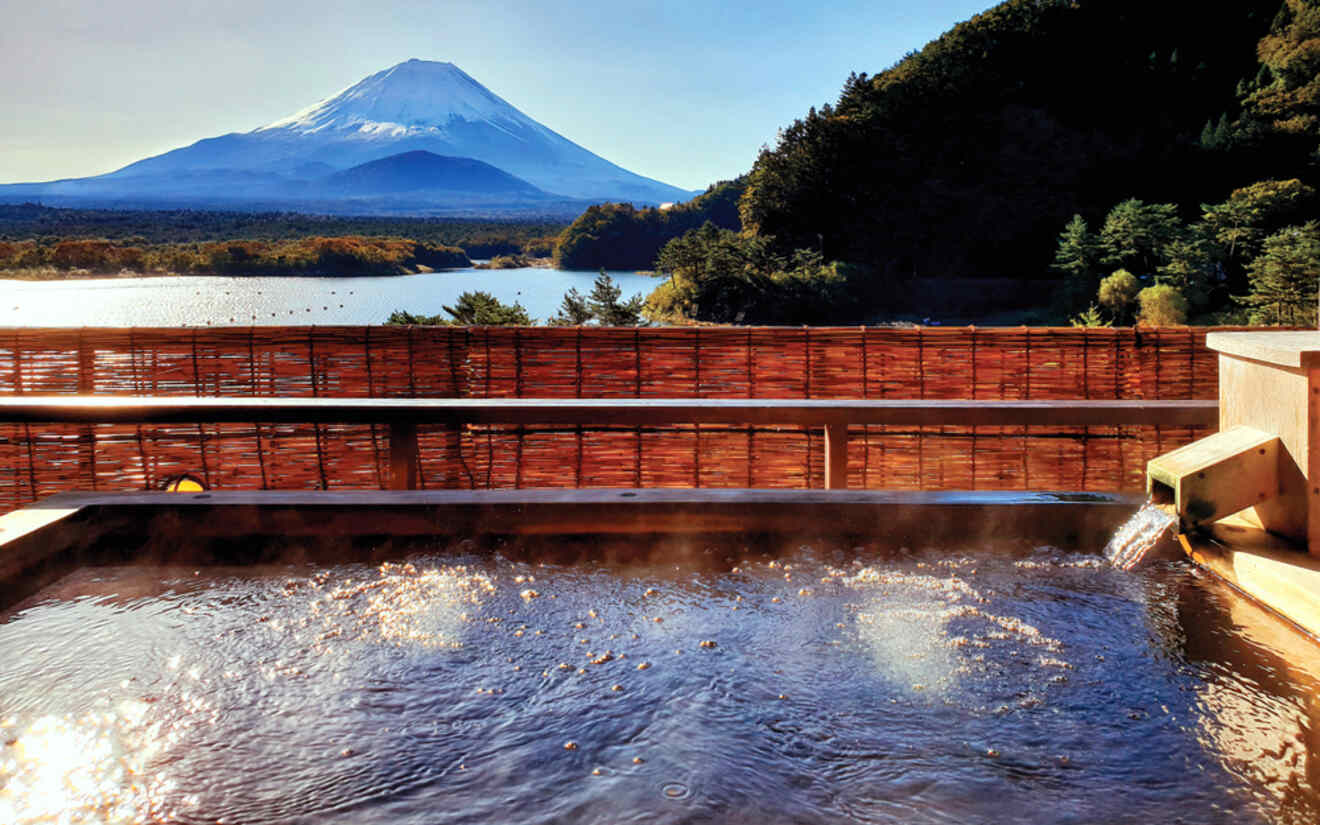 A serene view of Mount Fuji from a hot spring bath with a bamboo screen and a lake in the background.