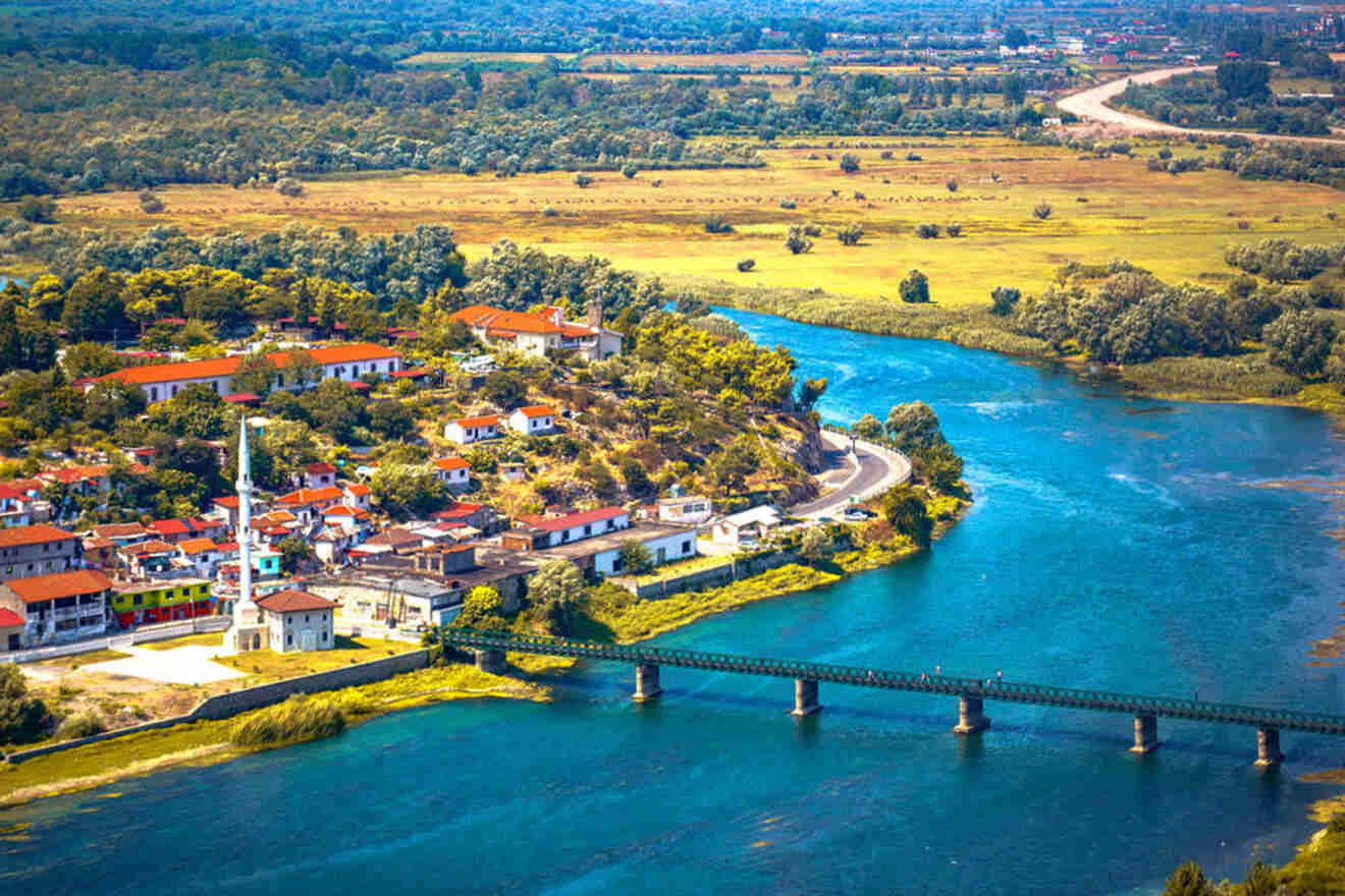 Aerial view of a vibrant village with red-roofed buildings, a river flowing alongside, a green bridge, and vast fields in the background.