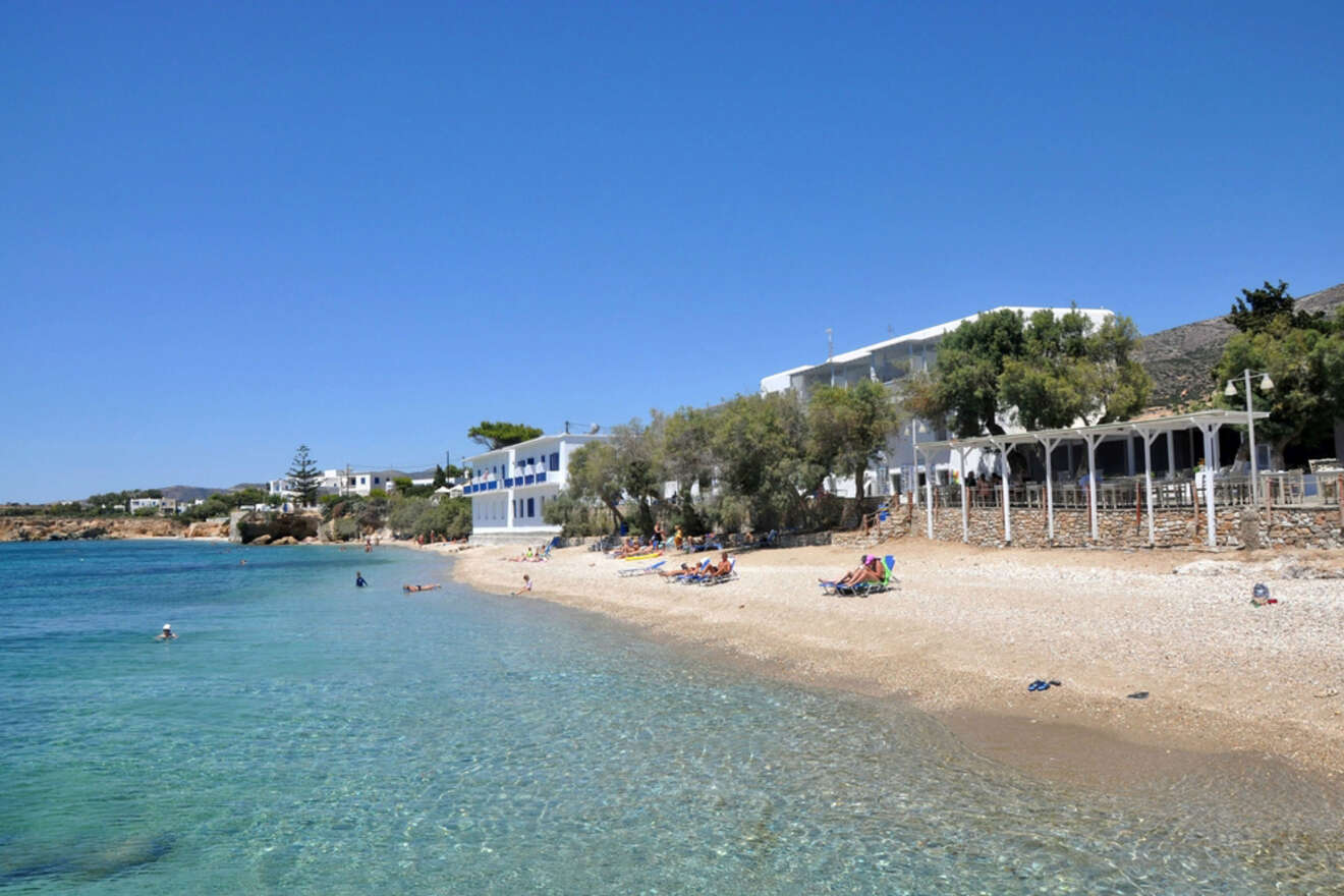 Clear waters of a beach with a scattering of people sunbathing and swimming, adjacent to a row of white buildings bordered by trees under a clear blue sky.