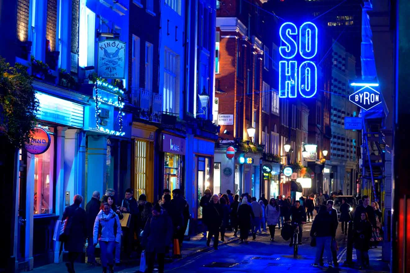 A crowd of people walking along a brightly lit street at night in Soho, London, with blue neon signs and colorful storefronts.
