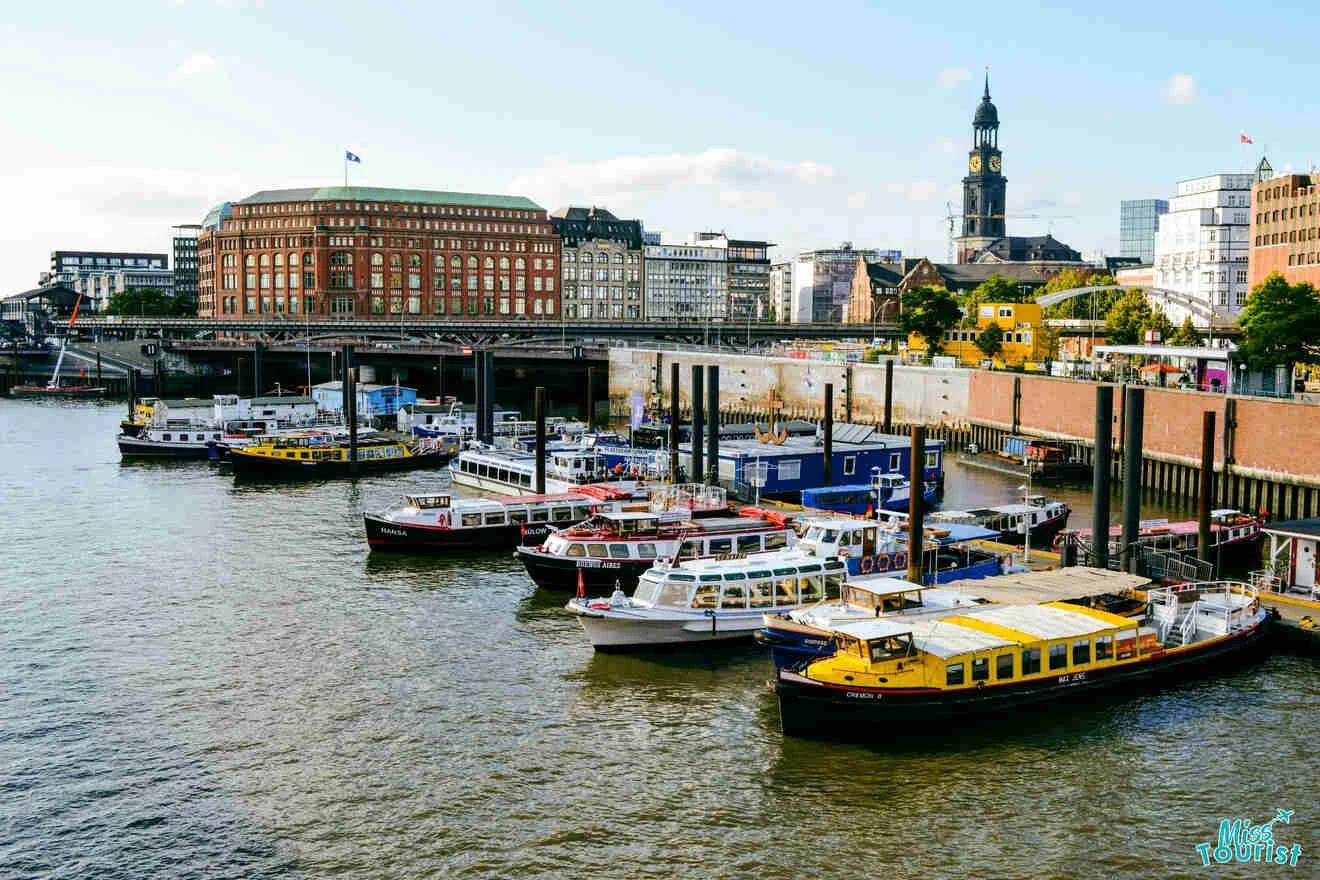 A lively dock with several boats moored at a harbor. Tall buildings and a clock tower are visible in the background under a clear sky.