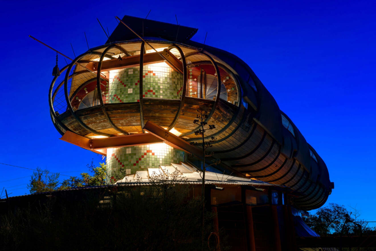 A uniquely shaped house illuminated at night, featuring a cylindrical structure with wood, tiles, and glass elements against a deep blue sky.
