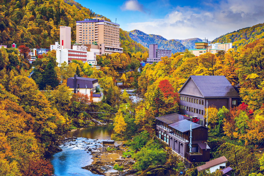 A scenic view of Jozankei Onsen, showcasing buildings nestled among colorful autumn trees and a flowing river.