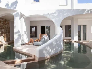 A person lies on a lounge chair by an arched white building with a surrounding pool of water, partially shaded by trees.